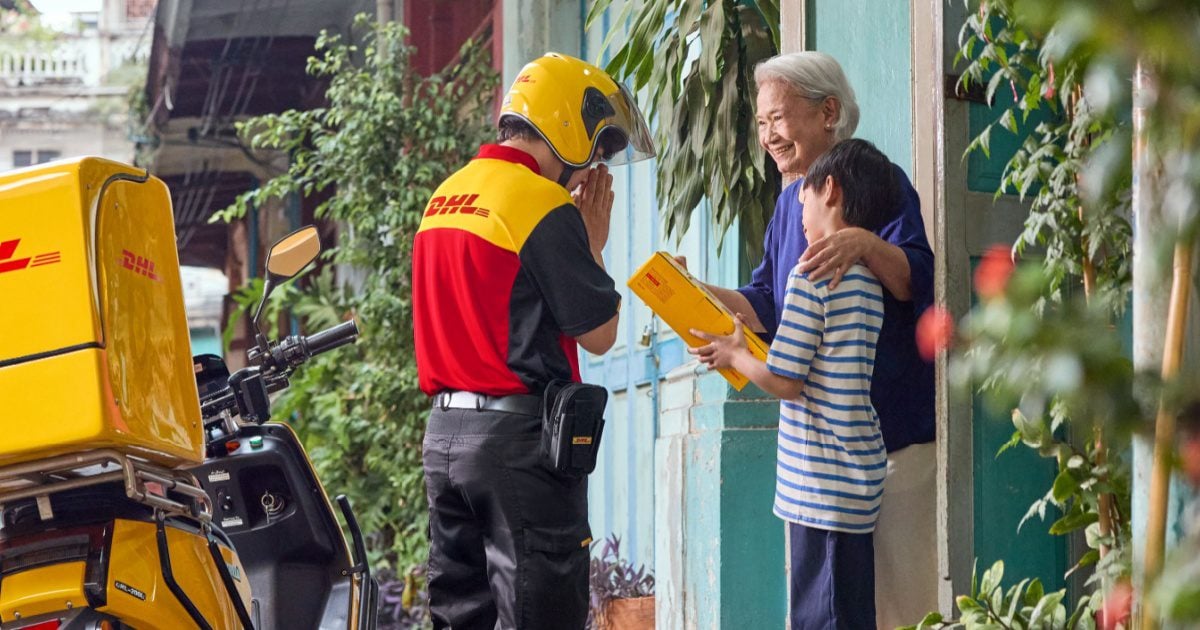 A DHL delivery person wearing a motorcycle helmet places his hands together in a Thai "Wai" greeting as a grandmother and her grandson receives a package.